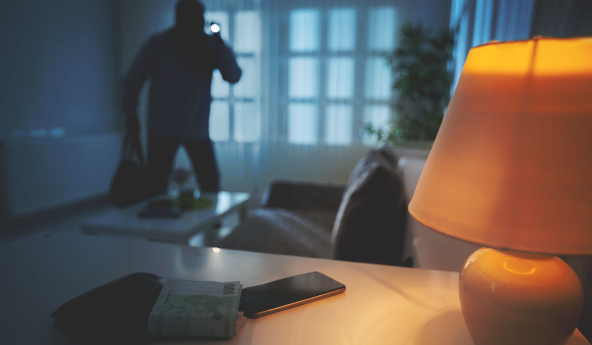 Dimly lit room with lamp and phone in foreground and blurred burglar in the background snooping around
