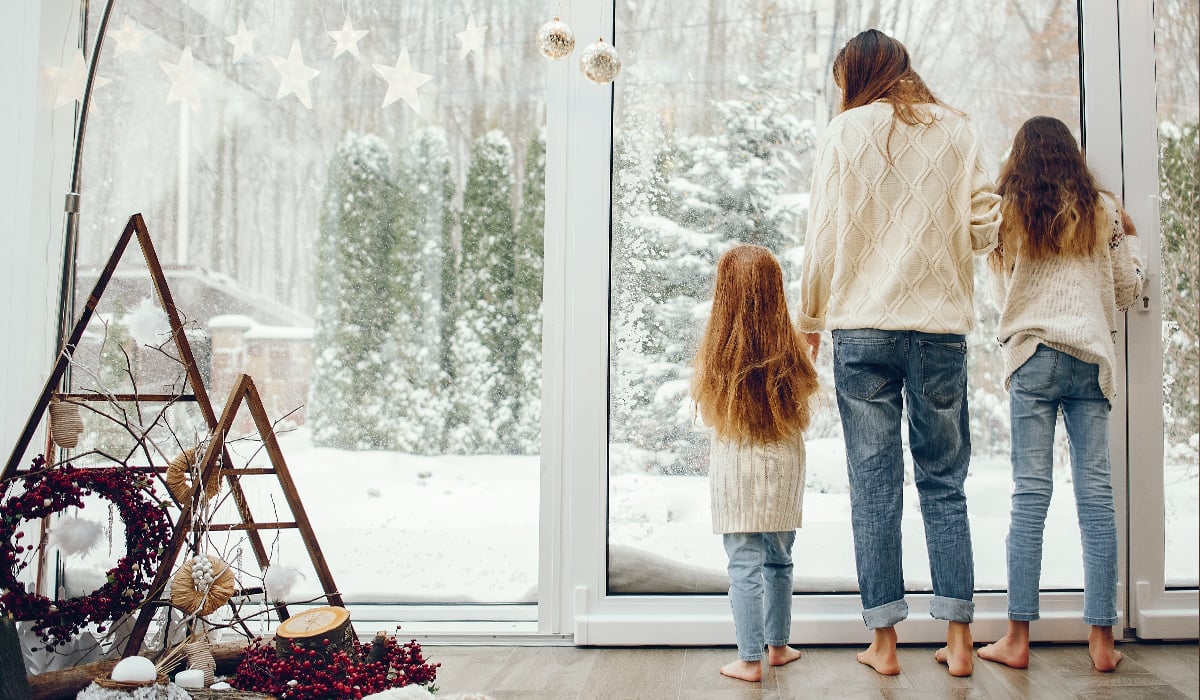 Mother and two daughters looking out snowy big glass window with Christmas decorations in foreground